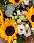 Prim-plan al florilor din buchetul "Buchet cu floarea-soarelui "Summer Vibes"", varianta Premium, evidentiind combinatia armonioasa de 7 flori de floarea-soarelui, 7 flori de lisianthus alb, 5 fire de limonium completate de 5 crengute de eucalipt si finisate cu 2 ambalaje decorative, o felicitare si 2 panglici elegante.