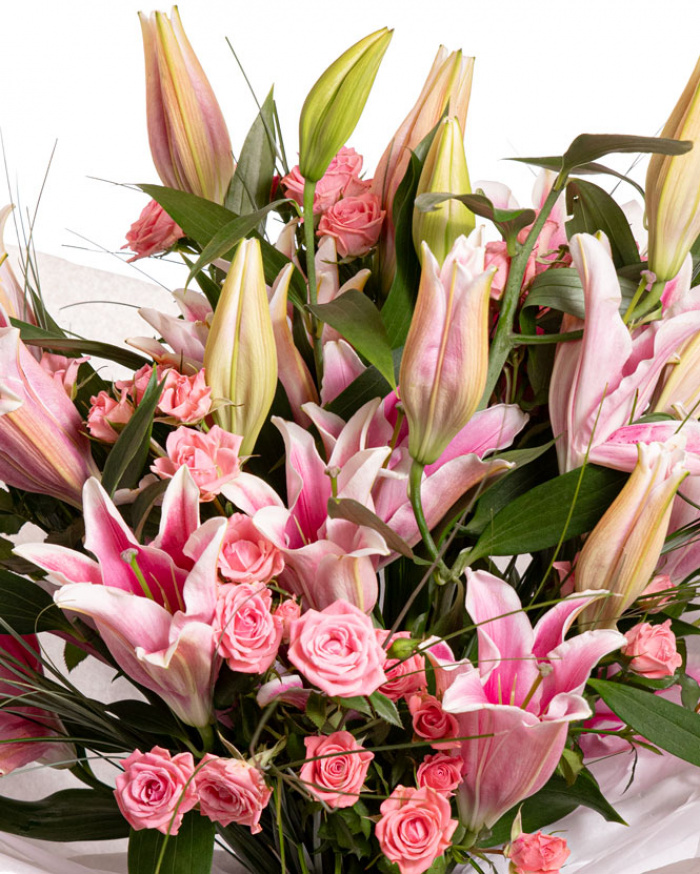 Prim-plan al florilor din buchetul "Buchet cu crini si trandafiri", varianta Premium, evidentiind combinatia armonioasa de 7 crini orientali roz, 8 trandafiri ramificati roz, completate de 20 de fire de beargrass si finisate cu 2 ambalaje decorative si 2 panglici inscriptionate elegante.