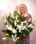 Perspectiva care evidentiaza proportiile si dimensiunea buchetului 'Buchet elegant de crini si trandafiri', varianta Premium, o compozitie bogata alcatuita din 7 crini orientali, 9 trandafiri albi, 5 frunze de monstera, 5 crengute de eucalipt si 7 crengute de ruscus mare, finisata elegant cu o panglica.