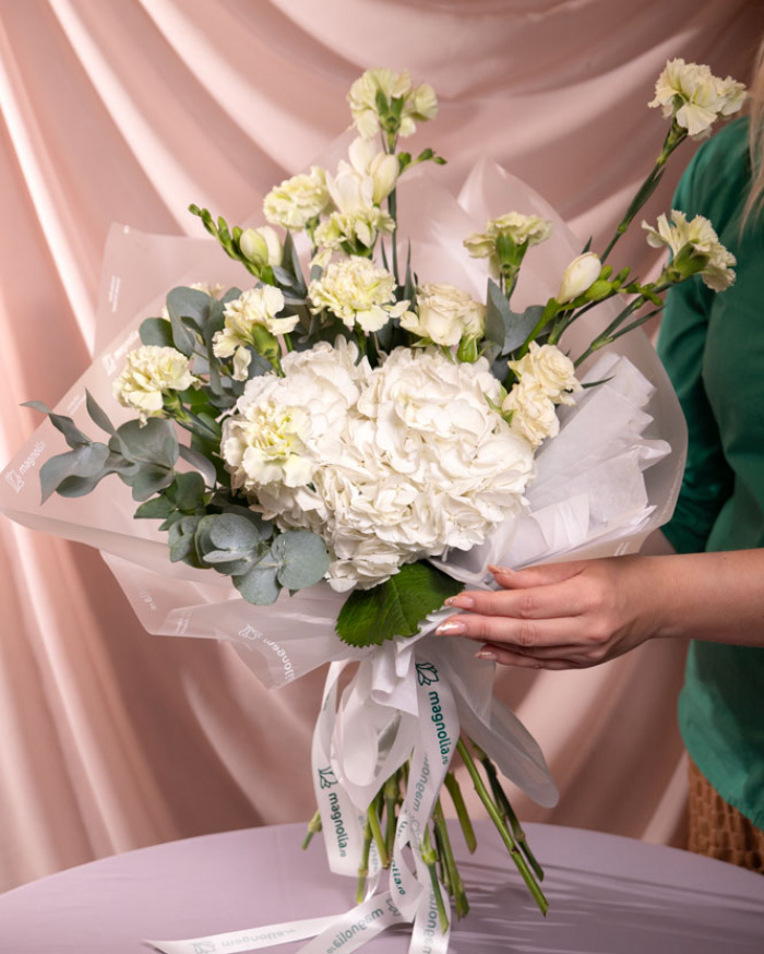 Perspectiva care evidențiază proporțiile și dimensiunea buchetului “Buchet cu flori albe și verzi”, varianta Premium, o compoziție bogată alcătuită din 1 Hydrangea albă, 3 Frezii albe, 10 Garoafe verzi, 3 Trandafiri ramificați albi și 2 crenguțe de Eucalipt, finisată elegant cu un ambalaj decorativ și o panglică inscripționată Magnolia.