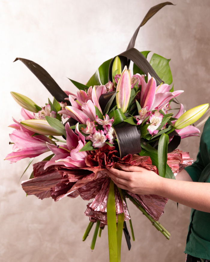 Perspectiva care evidentiaza proportiile si dimensiunea buchetului “Buchet crini orientali”, varianta Premium, o compozitie bogata alcatuita din 3 Crini orientali roz, 1 frunză de Monstera, 5 frunze de Cordiline, 4 flori de Alstroemeria, finisata elegant cu 1 element decorativ și o panglică neînscripționată.