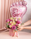 Perspectivă care evidențiază proporțiile și dimensiunea aranjamentului “Aranjament floral „It'a a Girl””, varianta Premium, o compoziție bogată alcătuită din 7 Trandafiri mov, 1 fir de Gypsophila, 2 flori de Eustoma somon, 2 crenguțe de Cinerea, finisată elegant cu 1 panglică neinscripționată, 1 perlă cu ac, ambalaj decorativ și 1 balon roz.