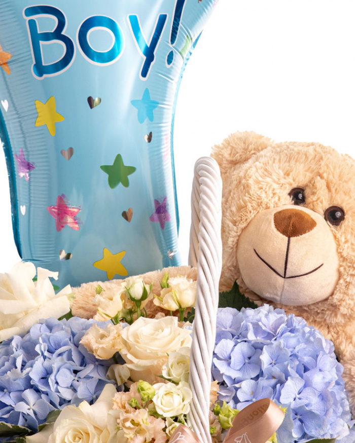 Prim-plan al florilor din aranjamentul “Aranjament floral nou-născut „Little Bear””, varianta Standard, evidențiind combinația armonioasă de 2 Hydrangea albastre, 3 Trandafiri albi, 2 flori de Eustoma somon și 2 Trandafiri ramificați albi, completate de 2 crenguțe de Pittosporum și finisate cu un coș mare, un urs maro mare, un balon festiv și 2 panglici inscripționate elegante.
