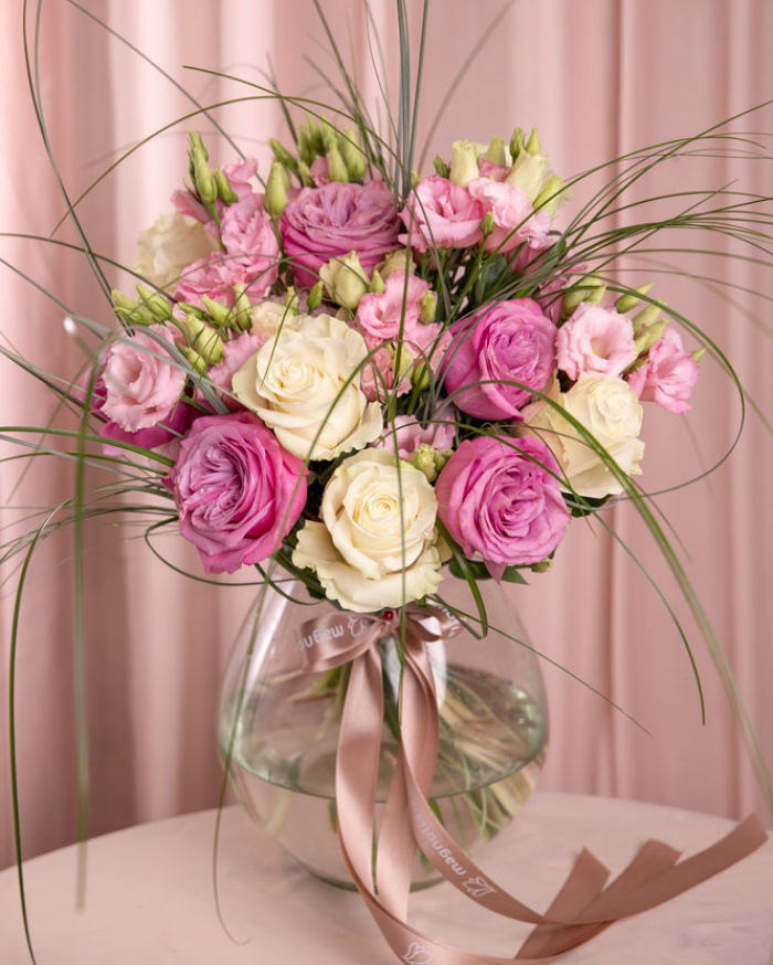Perspectivă care evidențiază proporțiile și dimensiunea buchetului “Buchet cu trandafiri mov și lisianthus”, varianta Premium, o compoziție bogată alcătuită din 5 Trandafiri albi, 6 Eustoma roz, 15 fire de Beargrass și 5 Trandafiri mov, finisată elegant cu 2 panglici inscripționate.