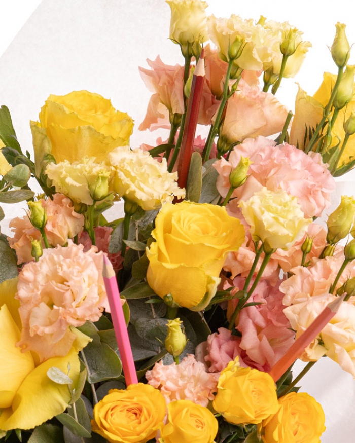Prim-plan al florilor din buchetul “Buchet trandafiri galbeni și creioane colorate „Back to school"”, varianta Premium, evidențiind combinația armonioasă de 5 Trandafiri galbeni, 3 Eustoma somon, 1 Trandafir ramificat galben și 1 set de creioane colorate, completate de 2 crenguțe de Eucalipt și finisate cu 1 ambalaj și 1 panglică elegante.
