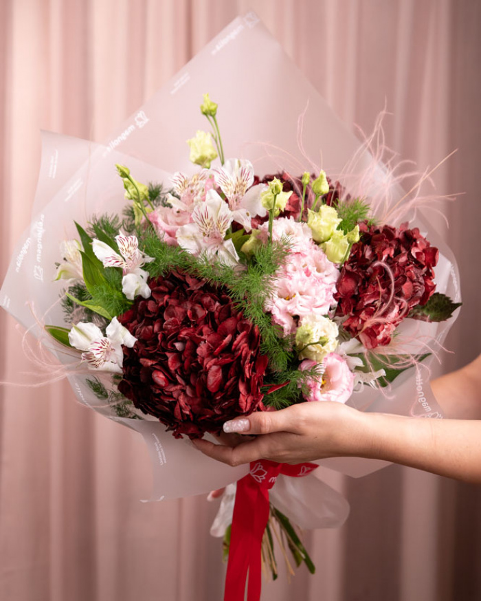 Perspectiva care evidentiaza proportiile si dimensiunea buchetului “Buchet cu Hortensii „Fall Elegance””, varianta Premium, o compozitie bogata alcatuita din 3 Hydrangea bordo, 2 Eustoma roz, 2 Alstroemeria, 4 fire de Asparagus umbelatus, 2 fire de Stypha, finisata elegant cu 1 ambalaj decorativ și 1 panglică inscripționată Magnolia.