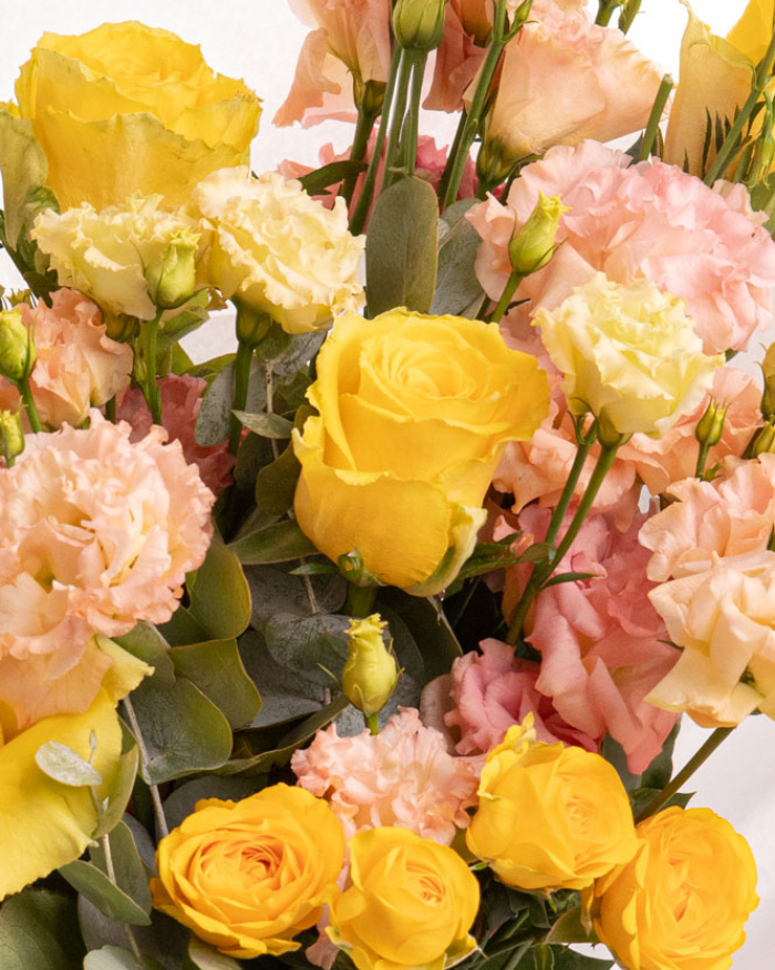 Prim-plan al florilor din buchetul “Buchet Trandafiri şi Eustoma”, varianta Premium, evidentiind combinatia armonioasa de 7 Trandafiri galbeni, 3 fire de Eustoma somon și 1 fir de Trandafir ramificat galben, completate de 2 crenguțe de Eucalipt si finisate cu ambalaj și o panglică elegante.
