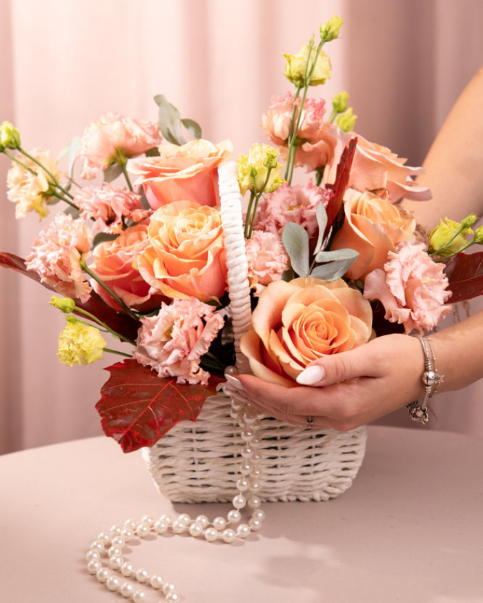Perspectiva care evidentiaza proportiile si dimensiunea coșului “Coș cu trandafiri portocalii și eustoma”, varianta Premium, o compozitie bogata alcatuita din 6 Trandafiri portocalii, 2 Eustoma somon, 1 ramură de Quercus, 1 fir de Asparagus cupru, 2 crenguțe de Eucalipt, finisata elegant cu 1 coș alb împletit și 1 șirag de mărgele.