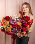 Perspectiva care evidențiază proporțiile și dimensiunea buchetului “Buchet de toamnă „Exotic Love””, varianta Premium, o compoziție bogată alcătuită din 5 Trandafiri roșii, 3 Leucospermum, 3 Brassica, 3 Carthamus, 2 Hydrangea burgundy, 1 Trandafir ramificat portocaliu, 2 Symphoricarpos, 3 fire de Asparagus cupru și 3 crenguțe de Quercus, finisată elegant cu ambalaj decorativ și o panglică Magnolia.